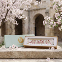 A rectangular ceramic dish with a pink floral pattern and gold trim, displayed in an open presentation box next to its mint green packaging box, set on a stone surface with cherry blossoms in the background.