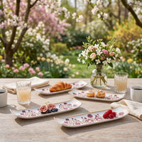 A styled outdoor dining scene on a wooden table in a garden setting, featuring several long rectangular ceramic plates with pink floral patterns and gold trim, alongside food items, glasses, and a vase of flowers.