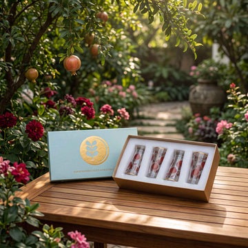 A gift set displayed on a wooden table in a lush garden setting. The set includes a light blue box with a gold circular logo, a matching lid, and three clear glass vials with pink and white patterned contents.