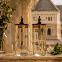 Two decorative glasses with gold patterns with wooden lids on a stone surface with a blurred architectural background.