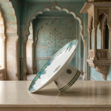 A white ceramic pedestal plate with blue and gold floral patterns, shown on a light stone surface with a blurred traditional architectural background.