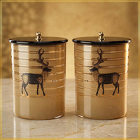 Two cylindrical decorative canisters with black lids and gold-toned bodies, each printed with a black deer motif, shown on a marble-like surface against a warm beige background.
