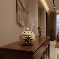A stoneware incense burner with a lid and palm tree motif sits on a wooden console table in a modern, warmly lit interior hallway.