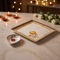 A square white ceramic plate with gold and purple borders, decorated with a traditional Diwali motif, is displayed on a marble table alongside a small matching heart-shaped dish. The background features warm bokeh lights from candles and flowers.