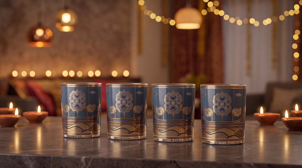 Four decorative blue and gold glass tumblers arranged in a row on a dark marble countertop, with warm bokeh lights and lit candles in the background.