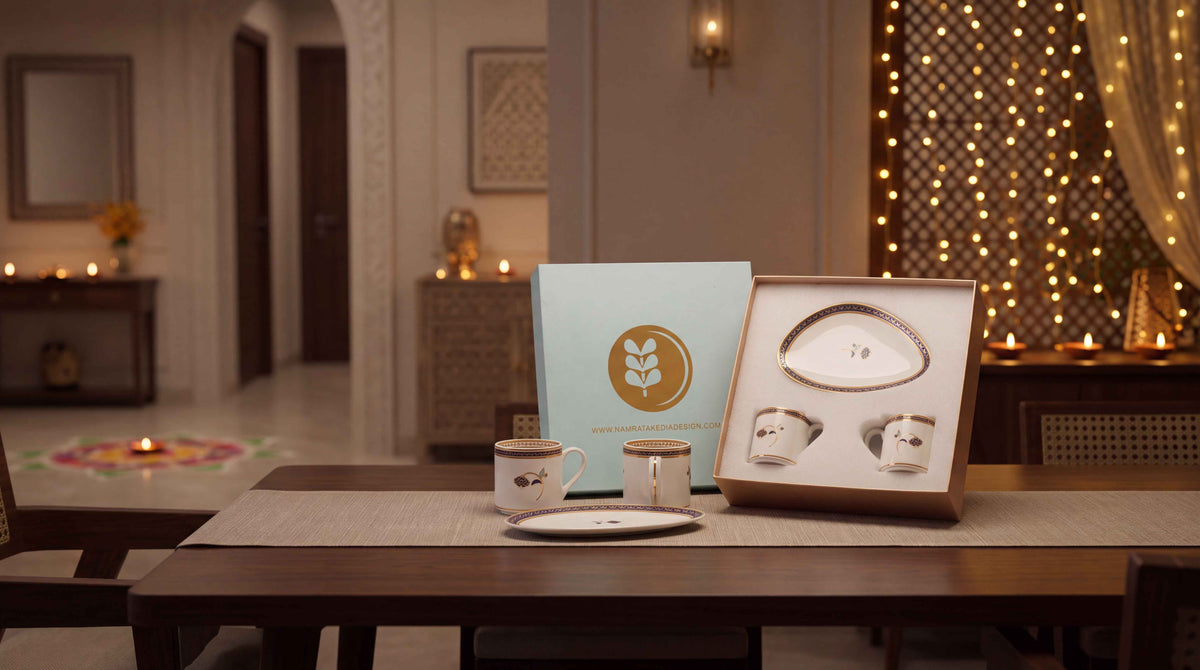 A boxed tea or coffee gift set displayed on a wooden table in a warmly lit indoor setting. The set includes a light blue outer box with NKD logo, an open box containing two white ceramic cups, a small white plate, and a matching saucer.