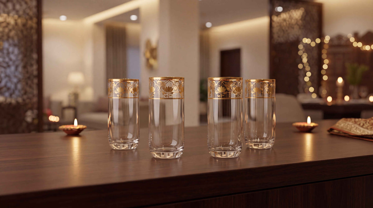 Four clear glass tumblers with ornate gold floral bands near the rims, arranged on a dark wooden table in a warmly lit indoor setting with candles in the background.