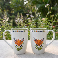Two white ceramic mugs with orange and green floral designs, displayed side-by-side on a light beige tablecloth with a blurred garden background.