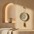 Decorative wall hanging with tassel on a beige wall with a shelf and vase.