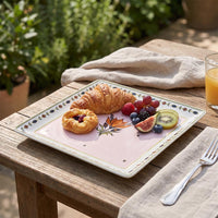 A square pink ceramic plate with a floral and leaf border design, styled on a wooden table outdoors with breakfast items including a croissant, fruit, a glass of orange juice, and a folded beige napkin.