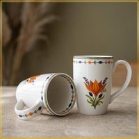 Two white ceramic mugs with colorful floral and geometric patterns, one standing upright and the other lying on its side to show the inside, set on a stone surface with a blurred background.