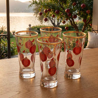 Four clear drinking glasses with red pomegranate illustrations and gold rims, arranged on a wooden table outdoors with a lake and tree in the background.