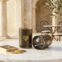 A decorative cylindrical shape glasses with a black diamond-patterned body and gold design, shown standing upright and lying on its side on a white marble table. A matching gold-toned coin or card box and several gold coins sit beside it.