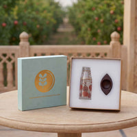 A gift set from Harvested by Design on a wooden table, featuring a sage green box with a gold leaf logo next to an open box containing a small glass bottle with red berries and a dark brown glass eye-shaped piece, with a blurred garden background.