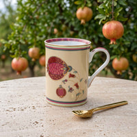 A cream-colored ceramic mug decorated with a red pomegranate and floral pattern, set on a stone table with a gold spoon in front, and pomegranate trees in the background.