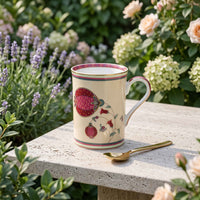 A cream-colored porcelain mug with a red pomegranate and floral design, resting on a stone garden bench with a gold spoon beside it and blooming flowers in the background.