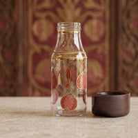 A clear glass bottle with a gold band and red pomegranate illustrations, shown next to a dark brown wooden cup on a stone surface with a patterned red and gold background.