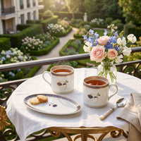 Two white ceramic tea cups with blue and gold floral detailing sit on a white tablecloth-covered table in a garden setting, alongside a small plate of cookies, a spoon, and a glass vase of pink and blue flowers.