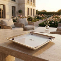 A square white ceramic serving platter with a gold floral border and small floral motifs, displayed on a stone table in an elegant outdoor patio setting.