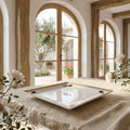 A square white ceramic serving platter with a gold-trimmed border and small dark floral motifs in the corners, displayed on a rustic stone table in a sunlit Mediterranean-style interior with arched windows.