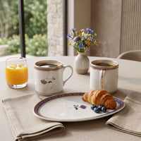 A breakfast table setting featuring a matching white ceramic plate and two mugs with blue and gold floral patterns, alongside a glass of orange juice, a small vase of flowers, and a croissant with blueberries.