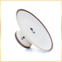 A white ceramic pedestal plate with a matching small bowl, decorated with blue and gold floral motifs and a blue-gold border, shown on a white background with a thin gold frame.