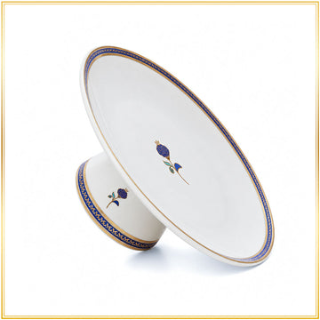 A white ceramic pedestal plate with a matching small bowl, decorated with blue and gold floral motifs and a blue-gold border, shown on a white background with a thin gold frame.