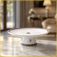 A white ceramic pedestal cake stand with blue and gold floral detailing, displayed on a polished marble table in a softly blurred, elegant interior setting.