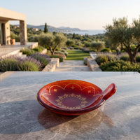 A small decorative red ceramic dish with gold geometric patterns sits on a gray stone table, with a luxurious outdoor garden and sea in the background.
