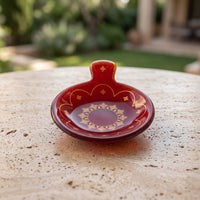 A small decorative red ceramic dish with a handle and gold geometric patterns, resting on a textured beige stone surface with a blurred outdoor garden background.