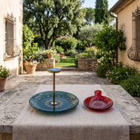 A single-tiered serving stand and matching plates displayed on a stone table in an outdoor garden courtyard. The stand features a teal blue top and bottom tier with gold decorative patterns and a central gold pole.