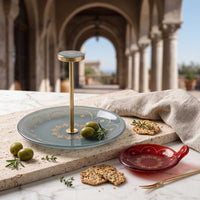 A decorative serving platter with a matching stand, styled on a stone ledge in an outdoor arched corridor. The platter is light blue with gold patterns, accompanied by green olives, crackers, a red bowl, and a beige cloth.