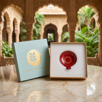 A red ceramic bowl with gold floral detailing presented in an open white box next to its light blue and gold branded packaging, set on a marble table with an ornate architectural background.