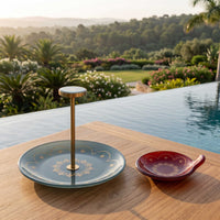 A decorative serving stand and matching small dish set on a wooden table beside an infinity pool, with a luxurious garden landscape in the background at sunset.