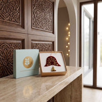 A decorative hanging ornament displayed in an open box next to its light blue gift box on a stone ledge. The ornament features a red and gold patterned bell-shaped fabric piece with a small gold bell hanging below, set against a background of carved wooden panels and warm string lights.