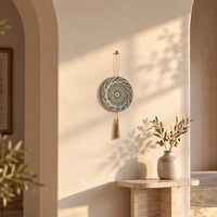 A circular woven wall hanging with a blue and turquoise spiral pattern and a tassel, displayed on a beige wall in a sunlit interior setting with a small vase and dried branches below.