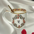 A decorated white porcelain coffee mug with gold and multicolor floral patterns, featuring a gold spoon, resting on a white silky surface with a few red strawberries nearby.