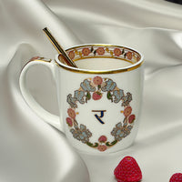 A decorated white porcelain coffee mug with gold and multicolor floral patterns, featuring a gold spoon, resting on a white silky surface with a few red strawberries nearby.