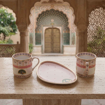 A matching set of two pink ceramic teacups with gold and multicolor patterns, and a matching pink teacup saucer, displayed on a stone table with a blurred traditional courtyard background.
