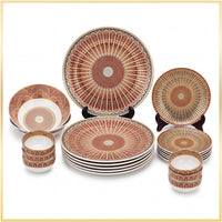 A coordinated dinnerware set with multiple plates and bowls arranged on a white background. The pieces feature a repeating sunburst or fan-like geometric pattern in warm terracotta, rust, beige, and muted blue-gray, with dark rim accents.