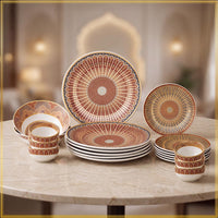 A coordinated dinnerware set arranged on a marble table in a softly blurred indoor setting. The set includes plates and bowls with a repeating fan-like geometric pattern in terracotta, beige, and muted blue, plus some pieces with thin black stripes.