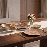A set of ceramic tableware including plates, bowls, and cups with a warm terracotta and beige sunburst pattern, arranged on a wooden dining table alongside gold cutlery and a vase of flowers.