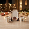 A ceramic tea or coffee set displayed on a table, featuring an oval plate on a stand in the center, flanked by two small cups. The set has a warm off-white/cream base with gold and reddish-brown decorative patterns, set against a softly blurred background with warm lights and candles.