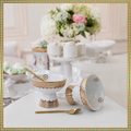 White porcelain dinnerware set with gold and silver patterns, including a pedestal bowl, a matching cup, a spoon, and a lid, arranged on a white table with a blurred elegant background.