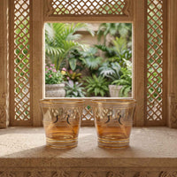Two clear glasses with gold decorative borders and black bird motifs, placed on a stone ledge in front of an ornate window with a garden view.