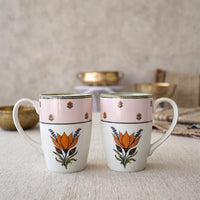Two white ceramic mugs with pink upper bands and orange floral designs, set on a textured beige surface with soft-focus decorative bowls in the background.