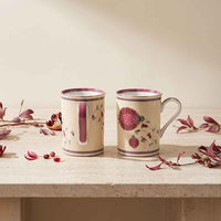 Two beige ceramic mugs with a pomegranate and floral design, displayed on a light stone surface with scattered dried pomegranate leaves.