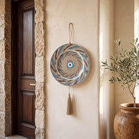 A circular woven wall hanging with a spiral evil eye pattern in blue, beige, and brown, suspended by a cord from a beige stucco wall next to a dark wooden door and a potted olive tree.