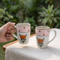 Two ceramic mugs with a pink top band and orange lotus flower design, one being held by a hand and one sitting on a table, with steam rising from both.