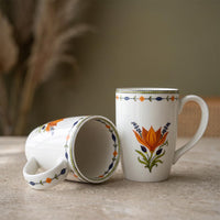 Two white ceramic mugs with colorful floral and geometric patterns, one standing upright and the other lying on its side to show the interior, set on a stone surface with a blurred background.